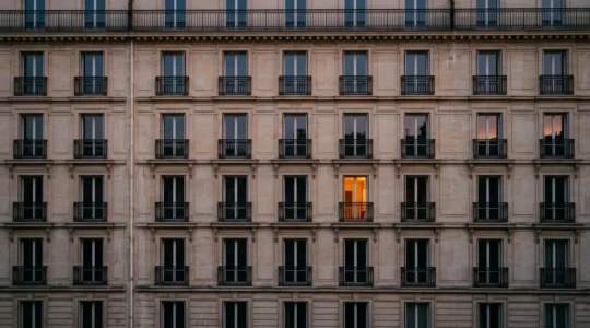Une fenêtre illuminée qui se distingue parmi une rangée uniforme de fenêtres sombres sur une façade haussmannienne parisienne, symbolisant un site qui capte l'attention sans changer de position dans les résultats de recherche.